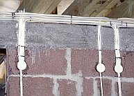 Pionowe i poziome kable elektryczne prowadzone wzdłuż szarej ściany z bloczków, przymocowane opaskami zaciskowymi, z okrągłymi puszkami instalacyjnymi w kolorze białym.